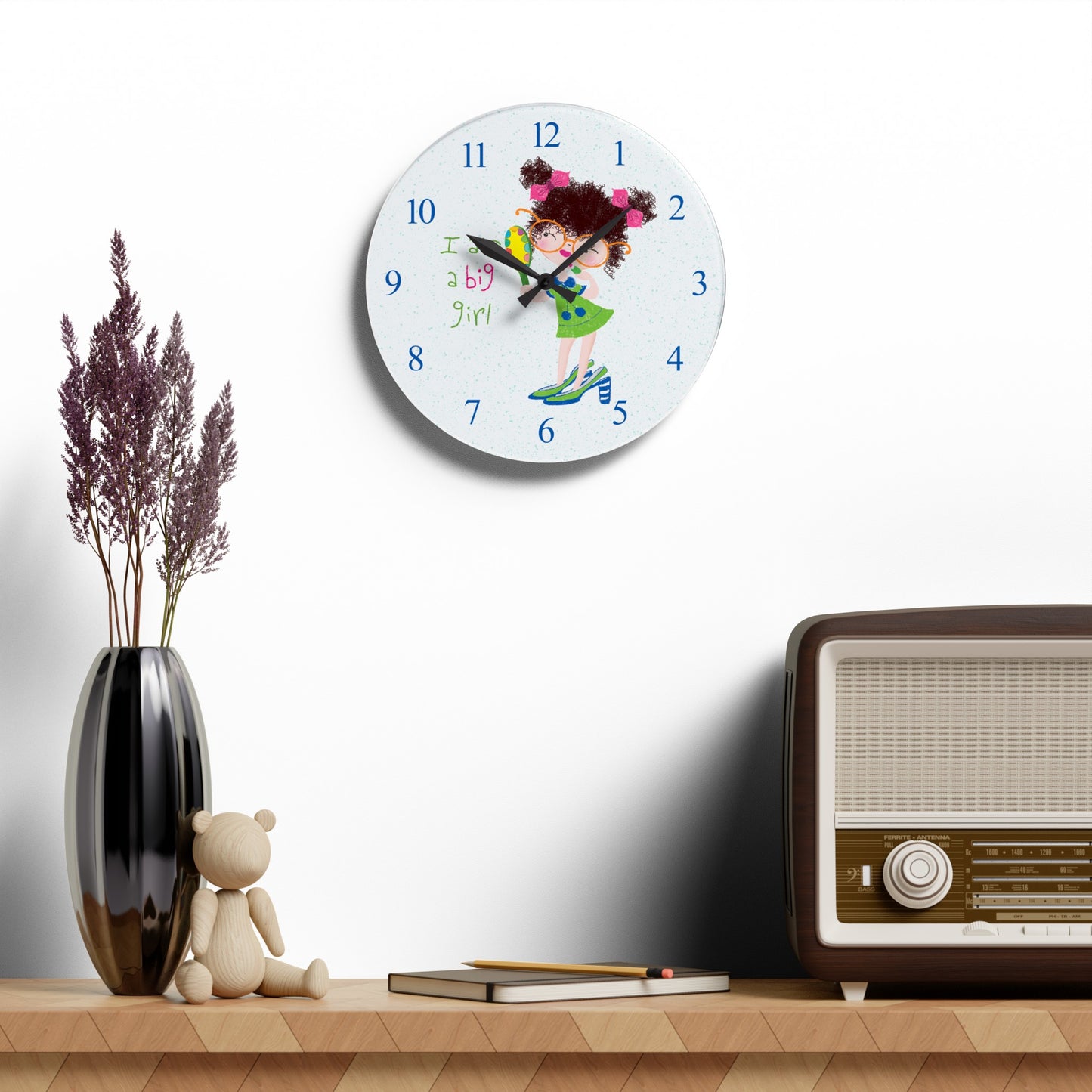 Humorous nursery acrylic wall clock hanging on a white wall featuring a cartoon girl with curly hair, oversized glasses, and a green dress, holding a mirror , with the text 'I am a big girl' alongside blue numbers on the clock face - on the wall, size 10.75”x10.75” (round ), Kidlic