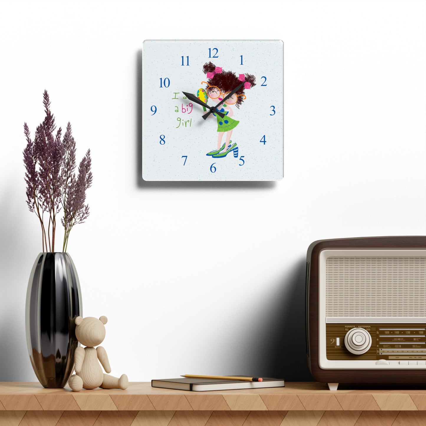 Humorous nursery acrylic wall clock hanging on a white wall featuring a cartoon girl with curly hair, oversized glasses, and a green dress, holding a mirror , with the text 'I am a big girl' alongside blue numbers on the clock face - on the wall, size 10.75”x10.75” (square ), Kidlic