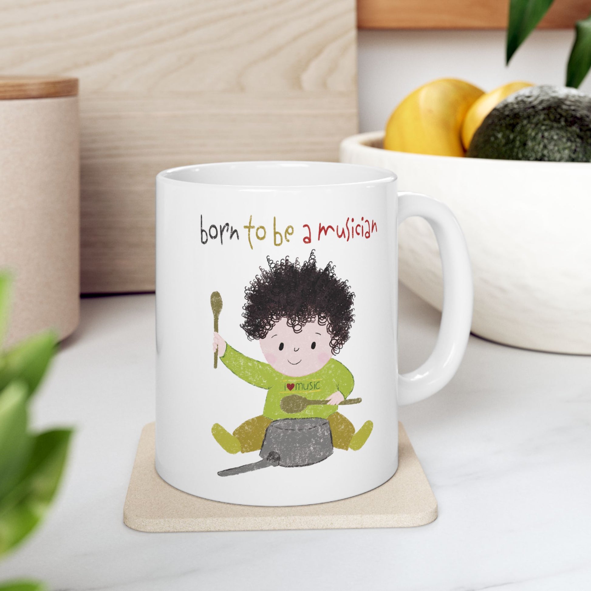 Humorous 11 oz nursery coffee mug featuring a cartoon baby boy playing a pot with a wooden spoon, with the text 'Born to be a musician’, on the kitchen table, kidlic