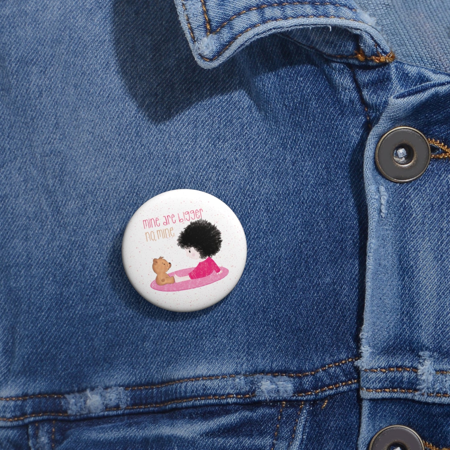 Playful nursery pin button featuring a cartoon child and a stuffed bear sitting on a pink mat, with the text 'mine are bigger,  no, mine’, size 1”, close-up, kidlic