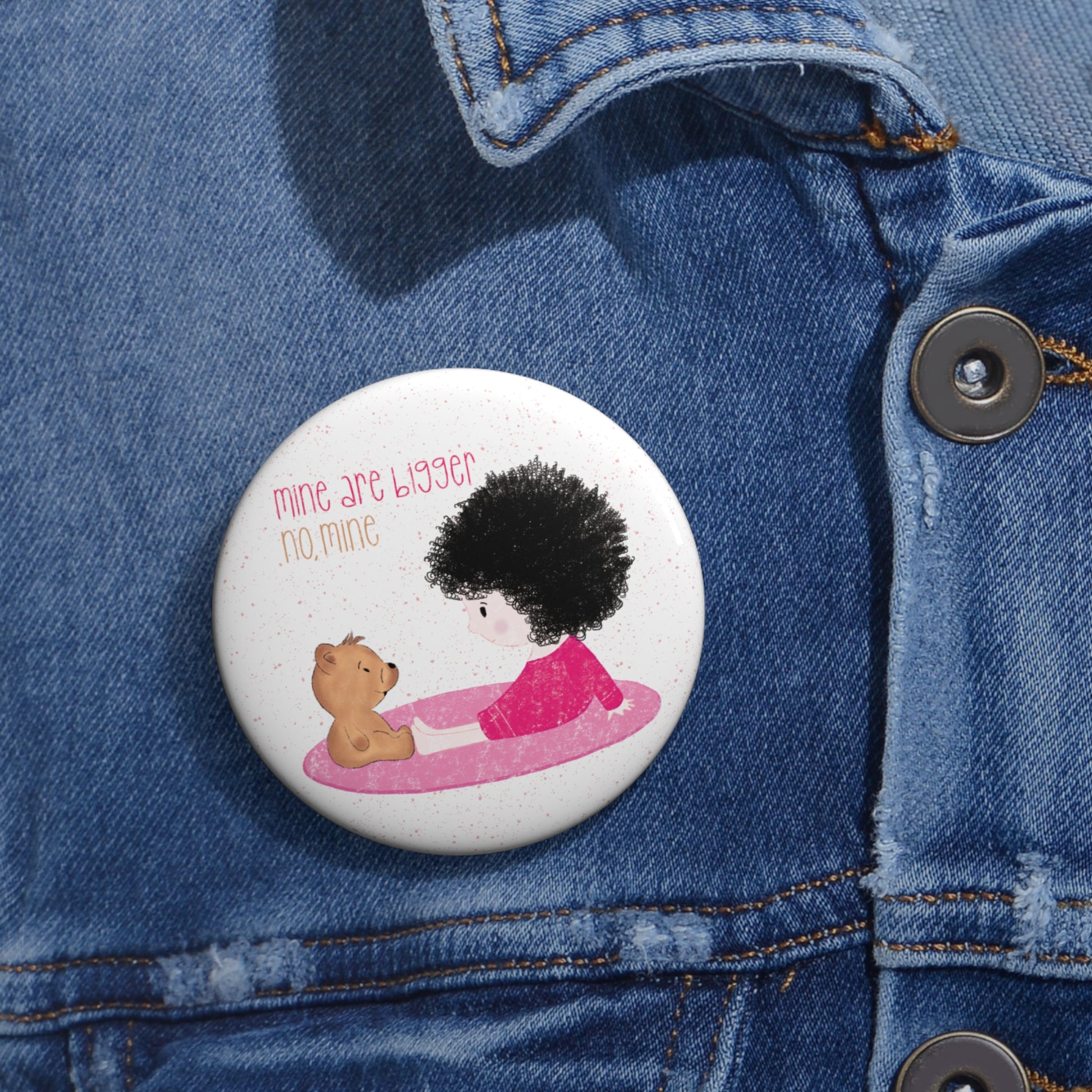 Playful nursery pin button featuring a cartoon child and a stuffed bear sitting on a pink mat, with the text 'mine are bigger,  no, mine’, size 2.25”, close-up, kidlic