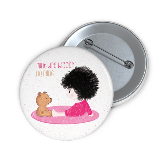 Playful nursery pin button featuring a cartoon child and a stuffed bear sitting on a pink mat, with the text 'mine are bigger,  no, mine’, size 3”, kidlic