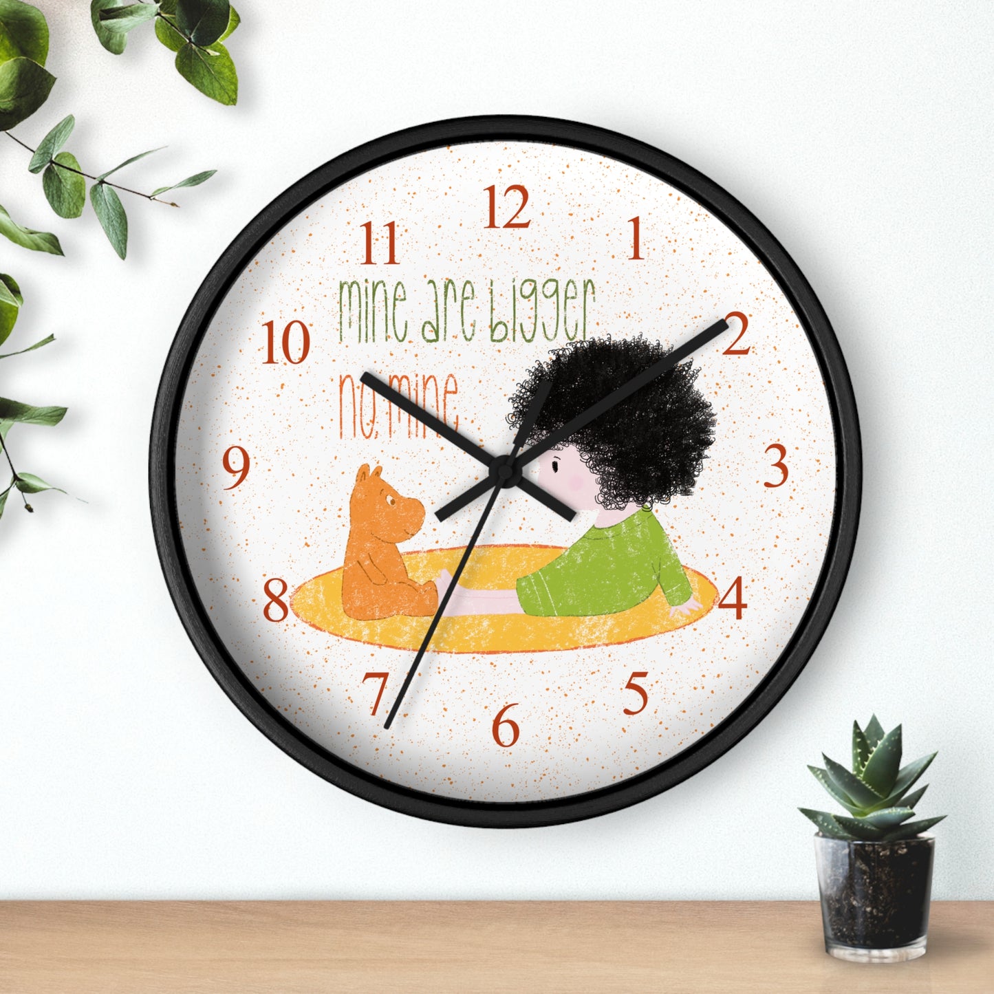 Funny 10” sized black round wall clock with black hands featuring a cartoon child sitting on a yellow mat, facing a stuffed bear, with the text 'mine are bigger' and 'no, mine,' and orange numbers on the clock face, kidlic