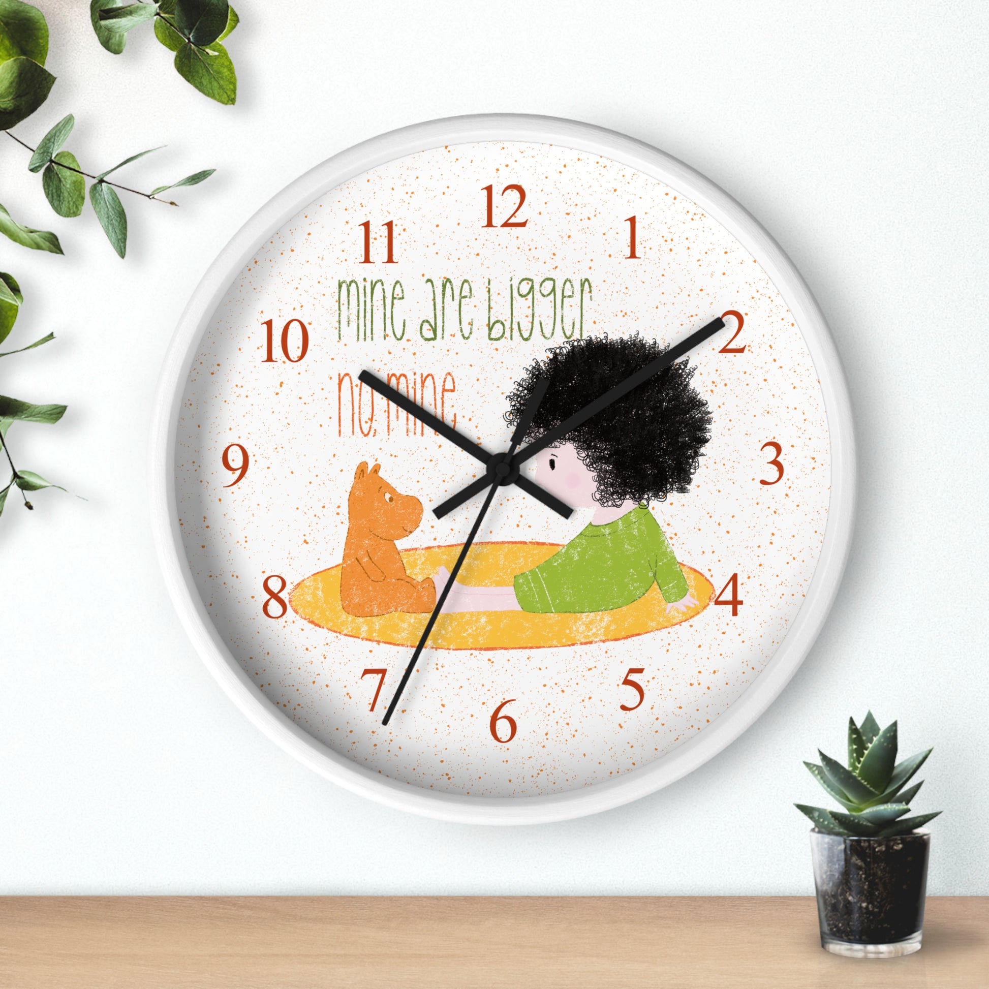 Humorous 10” sized white round wall clock with black hands featuring a cartoon child sitting on a yellow mat, facing a stuffed bear, with the text 'mine are bigger' and 'no, mine,' and orange numbers on the clock face, kidlic